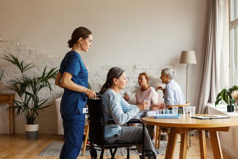Older adult welcomes family visitors in a bright assisted living lounge with chairs and a sign-in desk.